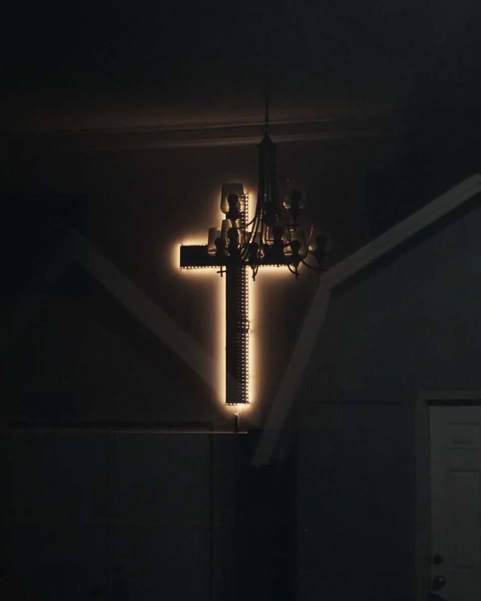 An illuminated cross on a dark wall beside a hanging chandelier