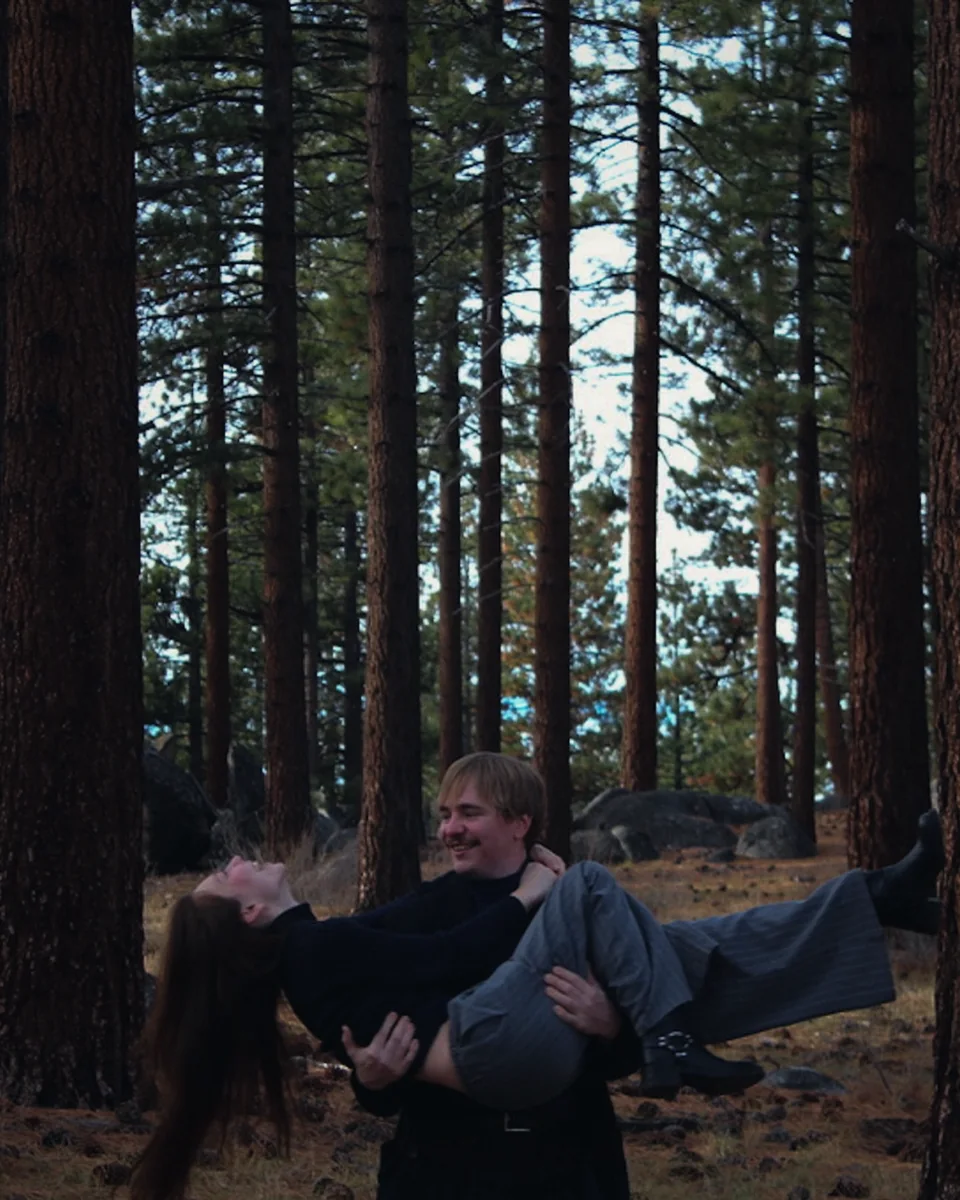 A man laughing while carrying a woman among tall pines